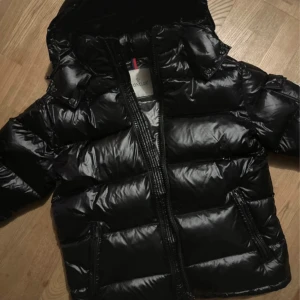 Svart Moncler dunjacka med huva - Säljer en svart, glansig dunjacka från Moncler med huva och dragkedja. Jackan har två fickor med dragkedja framtill och Moncler-logga på ärmen. Perfekt för kalla dagar och riktigt snygg streetstil.