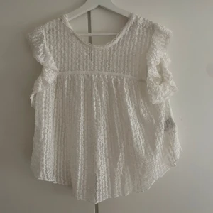 Vit blus med volangärmar - Supersöt vit blus med volangärmar och strukturmönster. Blusen har rund halsringning och är tillverkad i ett lätt, luftigt tyg som ger en romantisk känsla. Perfekt för dig som gillar en feminin och trendig stil.