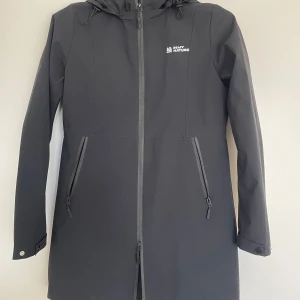 Svart regn & vind jacka från In My Nature - Svart regn och vind med huva från In My Nature. Jackan har långa ärmar, två dragkedjeförsedda fickor och justerbara muddar med tryckknappar. Vattentätt material och hel dragkedja framtill. Perfekt för blöta dagar och outdoor vibes.