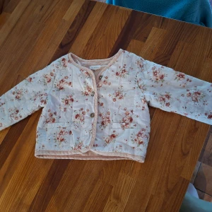 Blommig quiltad jacka från Newbie - Supersöt quiltad jacka från Newbie i storlek 86. Jackan är ljusbeige med blommigt mönster i rosa och bruna toner, har rundad hals, knappar framtill och två fickor. Perfekt för vår eller höst när det är lite kyligt ute.