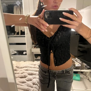 Svart ärmlös knytblus med spetsdetaljer - Säljer en svart ärmlös blus med v-ringning och knyt framtill. Toppen har ett genombrutet spetsliknande mönster och är croppad i modellen. Perfekt för dig som gillar trendiga och feminina detaljer.