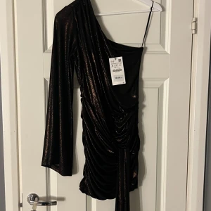 Svart glittrig oneshoulder klänning Zara - Supersnygg svart kortklänning från Zara med glittrigt tyg och draperad effekt. Klänningen har oneshoulder-design med en lång ärm och en axelbandslös sida. Perfekt för dig som vill sticka ut med en cool och trendig look.