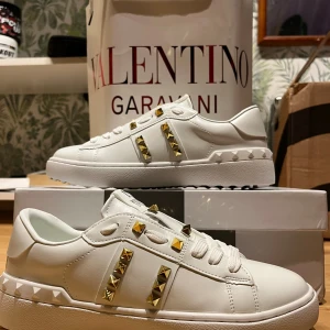Valentino Rockstud vita sneakers 38 - Säljer ett par vita Valentino Rockstud Untitled sneakers i skinn med ikoniska guldfärgade nitar på sidorna och runt hälen. Klassisk låg modell med snörning och chunky sula. Perfekta för dig som vill sticka ut med exklusiva detaljer och lyxig känsla.