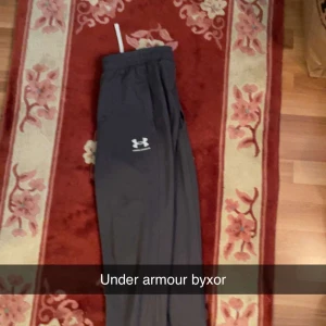 Svarta mjukisbyxor från Under Armour - Gråa mjukisbyxor från Under Armour med vit dragsko i midjan och logga på låret. Byxorna har en avslappnad passform och är perfekta för träning eller chill. Tillverkade i mjukt och bekvämt material.