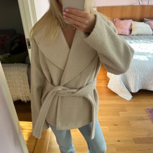 Beige omlottjacka från Vero Moda - Säljer en beige, mysig omlottjacka från Vero Moda med bälte i midjan och bred krage. Perfekt för lager-på-lager under höst och vinter. Använd få gånger!
