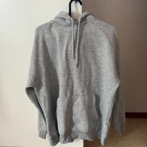 Grå hoodie med huva och magficka - En klassisk grå hoodie med huva och snörning samt en stor magficka framtill. Tröjan har ribbade muddar vid ärmslut och nederkant. Perfekt för chill dagar eller när du vill ha en enkel och stilren look.