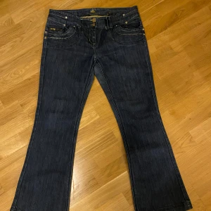 Mörkblå bootcut jeans River Island - Mörkblå jeans från River Island med bootcut passform. Snygga detaljer med kontrastsömmar, dubbla knappar och dragkedjor på fickorna både fram och bak. Klassisk femficksmodell i denim som sitter skönt och ger en cool look.
