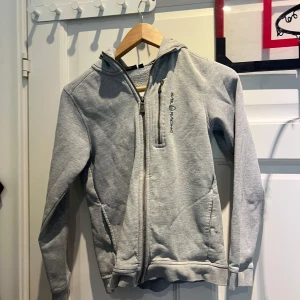 Grå hoodie från Sail Racing med huva - Snygg ljusgrå hoodie från Sail Racing med dragkedja hela vägen, broderad logga på bröstet och praktisk huva. Mjuk insida i fleece och två fickor framtill. Perfekt för dig som gillar en clean och sportig stil.
