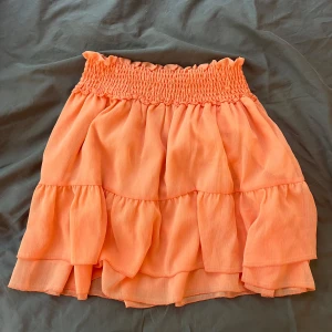 Volangkjol i orange från NA-KD - Supersöt kort volangkjol från NA-KD i en fräsch orange färg. Kjolen har smockad midja och flera lager med volanger som ger en flowy känsla. Perfekt för sommarens alla tillfällen och lätt att matcha med crop tops eller t-shirts.