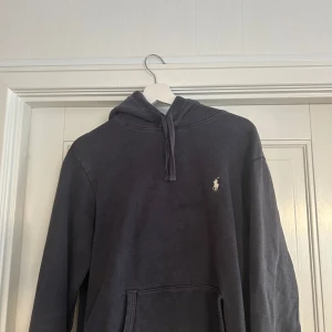 Mörkblå hoodie från Polo Ralph Lauren - Snygg mörkgrå hoodie från Polo Ralph Lauren med klassisk huva och snörning. Tröjan har en broderad vit logga på bröstet och en stor magficka framtill. Perfekt för dig som gillar stilrena och bekväma plagg med premiumkänsla. Storlek S passar även M. 