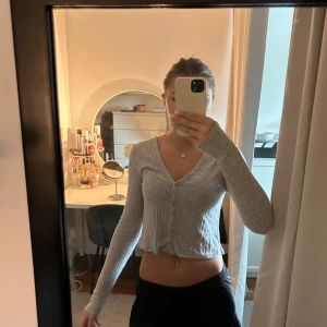 Grå croppad ribbad kofta - Säljer en ljusgrå croppad kofta med ribbad struktur och v-ringning. Koftan har långa ärmar och knappar framtill, perfekt att slänga över ett linne eller bära som den är. Enkel och stilren, passar till allt.