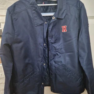 Marinblå vindjacka Tommy Hilfiger - Snygg marinblå vindjacka från Tommy Hilfiger med quiltat foder och klassisk krage. Jackan har tryckknappar, två fickor framtill och ett rött H-märke på bröstet. Perfekt för dig som gillar stilren och sportig look. Storlek 14y(164cm)