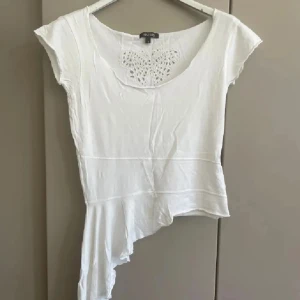 Vit asymmetrisk topp från Sisters - Superfin vit topp från Sisters med korta ärmar och unik asymmetrisk nederkant. Framtill finns en broderad fjäril i spets som ger en söt detalj. Tunt och mjukt bomullsmaterial, perfekt för dig som gillar en lite annorlunda look.