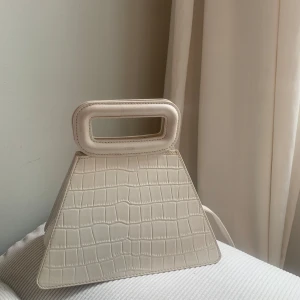 Beige handväska från Maje med krokoprägling - Superstylish beige handväska från Maje i unik trapetsform med croco-mönstrad yta. Väskan har ett rektangulärt handtag och avtagbar axelrem. Perfekt för dig som vill sticka ut med en trendig accessoar.