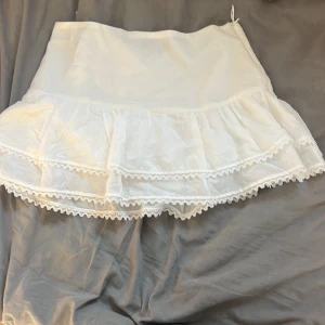 Vit volangkjol med spetsdetaljer - Supersöt vit kortkjol med dubbla volanger och fina spetsdetaljer längs kanterna. Kjolen har en romantisk och luftig känsla, perfekt för dig som gillar en feminin stil. Passar till sneakers eller boots för en trendig look.