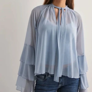 Ljusblå volangblus med knytband - Superfin ljusblå blus i transparent material med volangärmar och knytband vid halsen. Luftig och flowy passform, perfekt till jeans eller kjol. Ärmarna har flera lager volanger som ger en cool och trendig look.