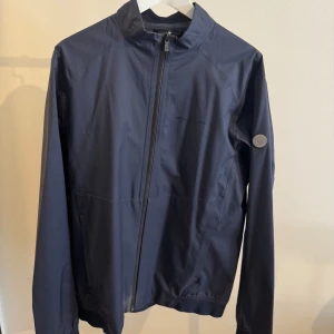 Blå vindjacka Sail Racing - Snygg marinblå vindjacka från Sail Racing med minimalistisk design och diskret logga på bröstet och ryggen. Jackan har hel dragkedja framtill, hög krage och långa ärmar. Perfekt för blåsiga dagar och enkel att matcha med det mesta.