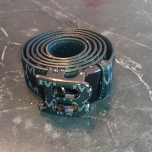 Svart skärp med B-spänne och M-mönst - Snyggt svart bälte i skinn med präglat M-mönster längs hela bandet och ett stort, glansigt B-format spänne. Bältet har en modern look och passar perfekt till jeans eller kostymbyxor. Materialet är slitstarkt och bältet har en stilren, urban känsla.