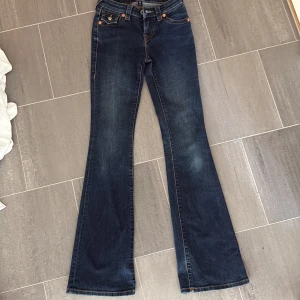 True Religion Becca Bootcut Jeans - Snygga mörkblå bootcut jeans från True Religion, modell Becca med medelhög midja. Klassiska kontrastsömmar, coola fickdetaljer med knappar och broderi bak. Tillverkade i stretchig bomullsdenim för en bekväm passform.