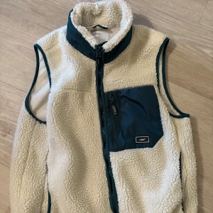 Vit fleeceväst från Bershka - Mysig vit fleeceväst från Bershka med hög krage och gröna detaljer. Västen har en stor grön bröstficka med dragkedja och matchande gröna kantband. Perfekt för lager-på-lager och chill dagar.