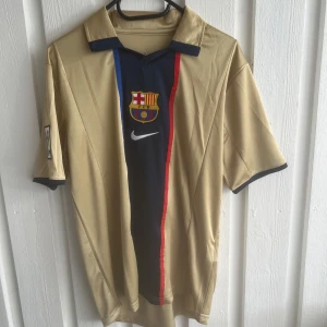 FC Barcelona Xavi 6 Nike fotbollströja - Snygg FC Barcelona fotbollströja i guldbeige med mörkblå och röda detaljer. Framsidan har klubbmärke, Nike-logga och en vertikal rand. Baksidan har XAVI och nummer 6 tryckt. Klassisk krage och korta ärmar. Perfekt för fans av Barca och Xavi.
