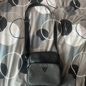 Svart axelväska från Guess med logomönster - Säljer en svart axelväska från Guess med justerbart band och klassiskt logomönster över hela väskan. Väskan har två stora fack med dragkedja och en fyrkantig form. Perfekt för att bära med sig allt viktigt och ändå se stilren ut.