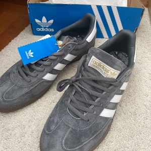 Adidas Handball Spezial grå/vit sneakers - Oanvända Adidas Handball Spezial sneakers i snygg kolgrå mocka med vita klassiska stripes och gummisula. Guldiga detaljer på sidan och tungan. Skorna har rund tå, snörning och retrovibe. Perfekta för dig som gillar streetstyle och ikoniska silhuetter. Säljer pga att de är för små. Sitter nog snarare som 44,5/45 än 45 1/3.