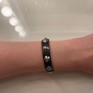 Svart läderarmband med nitar - Coolt svart armband i läder med silvriga, konformade nitar längs hela bandet. Justerbart spänne i metall för önskad storlek.
