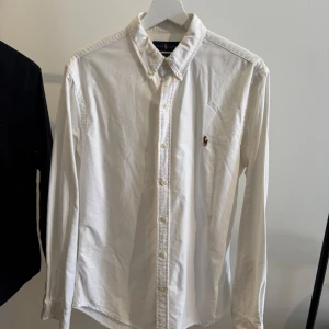 Vit skjorta från Ralph Lauren - Klassisk vit skjorta från Ralph Lauren med button-down krage och broderad logga på bröstet. Skjortan har lång ärm, knappar framtill och är tillverkad i mjuk bomull. Perfekt för en clean och stilren look.