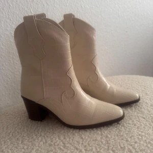 Beiga cowboy boots med krokoprägling - Snygga beige cowboy boots med krokodilpräglat mönster och markerad klack. Skorna har spetsig tå och är tillverkade i syntetmaterial med cool western-inspirerad design. Perfekta för dig som vill sticka ut med trendiga boots i ljus ton. Använd en gång.