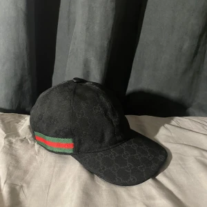 Svart keps från Gucci med GG-mönster - Snygg svart keps från Gucci med diskret GG-monogram över hela kepsen. Klassisk böjd skärm och justerbar rem baktill i skinn. På sidan finns den ikoniska gröna och röda randen. Perfekt accessoar för att lyfta din outfit.