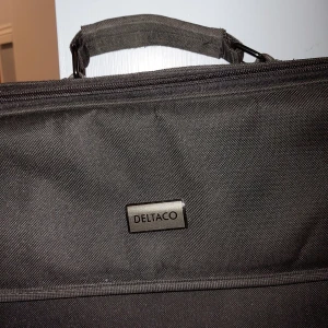 Svart datorväska från Deltaco - En stilren svart datorväska från Deltaco med axelrem och handtag. Väskan har flera fack med dragkedja, perfekt för laptop och tillbehör. Tillverkad i slitstark polyester och har en enkel, clean look som funkar till skolan eller jobbet.