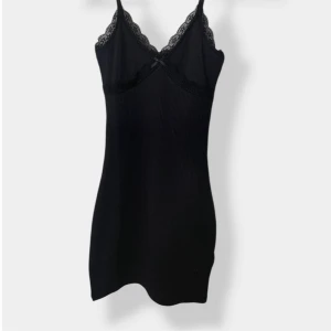 Svart ribbad spetsklänning med rosett - Supersöt svart kortklänning med ribbad struktur, smala axelband och v-ringning. Spetsdetaljer längs urringningen och en liten rosett framtill ger en extra touch. Perfekt för dig som gillar en feminin och stilren look.