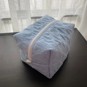 Rutig blå och vit necessär i bomull - Söt necessär i blåvitt rutigt bomullstyg med vit dragkedja. Insidan är fodrad med vitt tyg och små blå blommor. Perfekt för smink eller småprylar, och den boxiga formen gör den rymlig och lätt att packa ner i väskan.                        Bredd: 12 Längd: 18  Höjd: 12