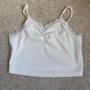 Vit ribbad linnetopp med spets - Supersöt vit ribbad linnetopp med tunna axelband och spetsdetaljer längs urringningen och ryggen. Toppen har en lätt rynkning framtill och är croppad i modellen. Perfekt att styla  med jeans eller kjol för en fräsch look. Storlek 158🥰