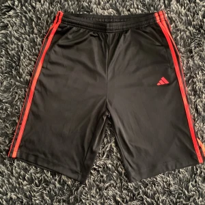 Svarta Adidas shorts med röda ränder - Svarta sportiga shorts från Adidas med röda ränder längs sidorna och klassisk Adidas-logga framtill. Elastisk midja för skön passform. Perfekta för träning eller fotboll, tillverkade i lätt polyester som andas. Passformen är bra och stämmer med den givna storleken.