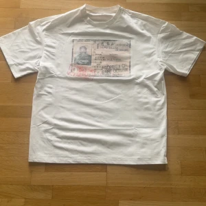 Vit t-shirt Dirty South Art Gallery - Vit t-shirt från Dirty South med tryck framtill som liknar ett ID-kort och stort grafiskt tryck på ryggen med text och siffror i svart och rött. Klassisk passform med rund hals och korta ärmar. Perfekt för dig som gillar streetwear och unika prints.