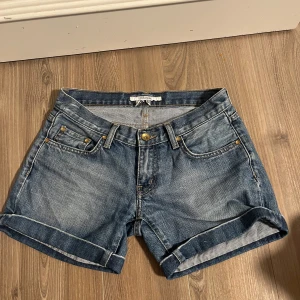 Blå y2k low waist jeansshorts från Zara - Snygga blå y2k low waist jeansshorts från Zara Basic med klassisk femficksdesign och uppvikta benslut. Shortsen har guldiga nitar och knappar samt dekorativa broderier på bakfickorna. Perfekta för varma dagar och enkel att matcha med allt.