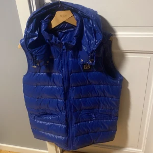 Blå dunväst med huva - Snygg blå dunväst med glansig finish och avtagbar huva. Västen har dragkedja framtill, två fickor med dragkedja och quiltad design. Perfekt för lager-på-lager och ger en sportig vibe till din outfit.