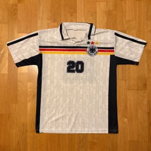 Tysklands retro fotbollströja Bierhoff - Snygg vit Tyskland fotbollströja med svarta, röda och gula ränder över bröstet, nummer 20 och Bierhoff på ryggen. Klassisk krage, korta ärmar och diskret mönster i tyget. Tysklands landslagsemblem med tre stjärnor på bröstet. Perfekt för fotbollsfans!