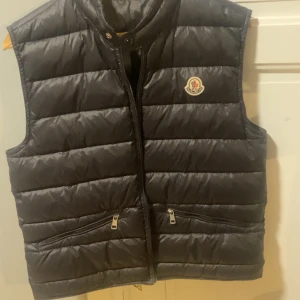 Svart dunväst från Moncler - Snygg svart dunväst från Moncler med klassisk quiltad design och logotyp på bröstet. Västen har två dragkedjefickor framtill och hög krage. Perfekt att slänga över en hoodie eller tröja för extra värme och stilpoäng. Liten skada i fickan detta syns ej och därför billiga priset