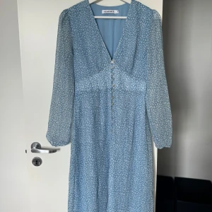 Blå mönstrad klänning från Adoore - Supersöt blå långklänning från Adoore med vitt leopardmönster, v-ringning och knappar framtill. Klänningen har långa, transparenta ärmar och en liten slits framtill. Perfekt för vår och sommar!