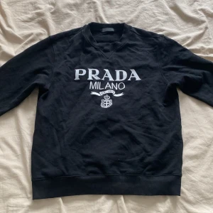 Prada Milano tröja - Säljer en svart sweatshirt från Prada Milano med vit broderad logga framtill. Tröjan har klassisk rund halsringning, långa ärmar och ribbade muddar. Perfekt för dig som gillar stilrena designer och vill ha något från ett lyxigt märke.