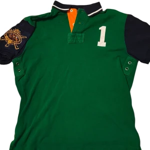 Grön pikétröja Polo Ralph Lauren M - Snygg grön pikétröja från Polo Ralph Lauren i custom fit. Tröjan har mörkblå ärmar, vit siffra 1 på bröstet, broderat emblem på ena ärmen och orange detalj vid knäppningen. Klassisk krage med vit rand och korta ärmar.