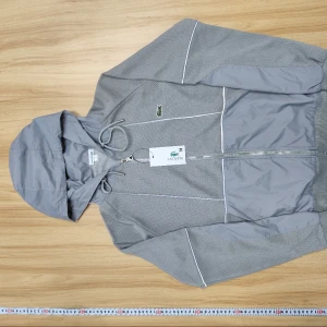 Lacoste set grå  - Snygga grå mjukisbyxor från Lacoste med elastisk midja och dragsko. Byxorna har diskret meshstruktur, muddar vid benslut och fickor med detaljer i blankare tyg. Klassisk Lacoste-logga på ena fickan. Perfekta för chill eller sport.