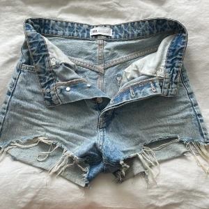 Jeansshorts från Zara - Snygga ljusblå jeansshorts från Zara, fransiga benslut. Klassisk femficksmodell med knappgylf och slitna detaljer. Perfekta till sommaren och lätta att matcha med allt.