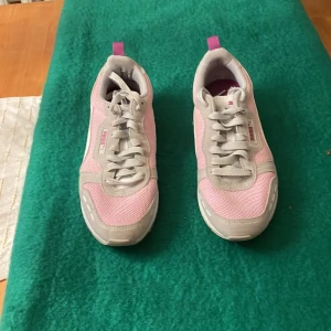 Rosa och grå sneakers från Puma - Snygga sneakers från Puma i rosa och grått med vita detaljer. Skorna har mesh- och syntetmaterial, klassisk snörning och en platt sula. Färgstarka detaljer i lila och persika på hälen ger dem en sportig vibe. Perfekta för dig som gillar färg och streetstyle.