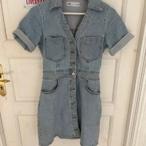Kort jeansklänning från Zara - Snygg ljusblå jeansklänning från Zara med kort ärm, knappar hela vägen framtill, två bröstfickor och två stora fickor vid höfterna. Klassisk krage och uppvikta ärmslut ger en cool vibe. Perfekt för dig som gillar denim!