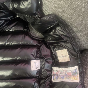 Svart dunväst från Moncler med huva - Säljer en svart, glansig dunväst från Moncler med avtagbar huva och dragkedja framtill. Västen har två fickor med dragkedja och klassisk Moncler-logga på bröstet. Perfekt för lager-på-lager och riktigt snygg till streetwear-stilen.