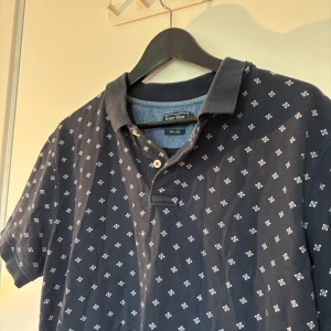 Mörkblå mönstrad piké från East West - Snygg mörkblå pikétröja från East West i slim fit-modell. T-shirten har korta ärmar, klassisk krage och ett diskret vitt rutmönster över hela plagget. Perfekt för dig som gillar stilrena men ändå unika detaljer. Materialet är mjukt och skönt, troligtvis bomull. Sitter som en M